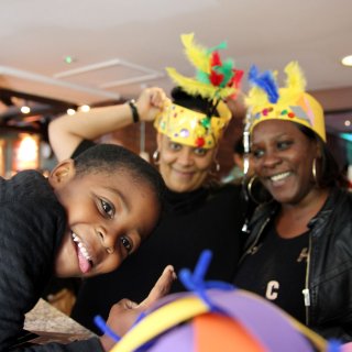 Photo of a child and two adults smiling to the camera at a paper hat making workshop at Watershed