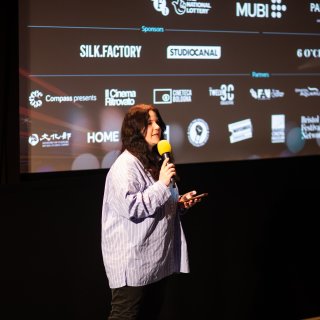 A woman speaks into a mic on stage in front of a cinema screen displaying sponsor and partner logos