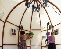 An image of the inside of a domed house with curved iron ribs onto which white canvas has been tautly wrapped around. Traditional Somali prints on the outside of the canvas are also visible from the inside of the house. We can see the backs of two women facing various colourful small woven structures, and a potted plant, all hanging from the ribs of the house. A microphone and lights are also visible inside the space.