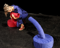 A person in a blue boilersuit is hugging a soft red tubular sculpture facing a similar looking blue sculpture. This is in a black box theatre stage.