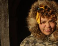 A women of colour squinting into the sun. She is wearing a green camouflage coat, with a fur trim around her face and her yellow hair tie  is visible through her curly afro.