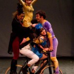 A tower of five circus performers - all balanced on a bike, with a woman in yellow on the top with her arms outstretched