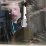 a blurred image taken through glass of Jez singing in a sound booth with a mic in front of him