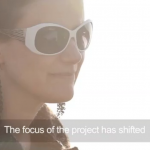 A screen grab of a captioned film. There is a woman's face, shot against the sun, she is wearing long earrings and sunglasses. The text says 'The focus of the project has shifted'