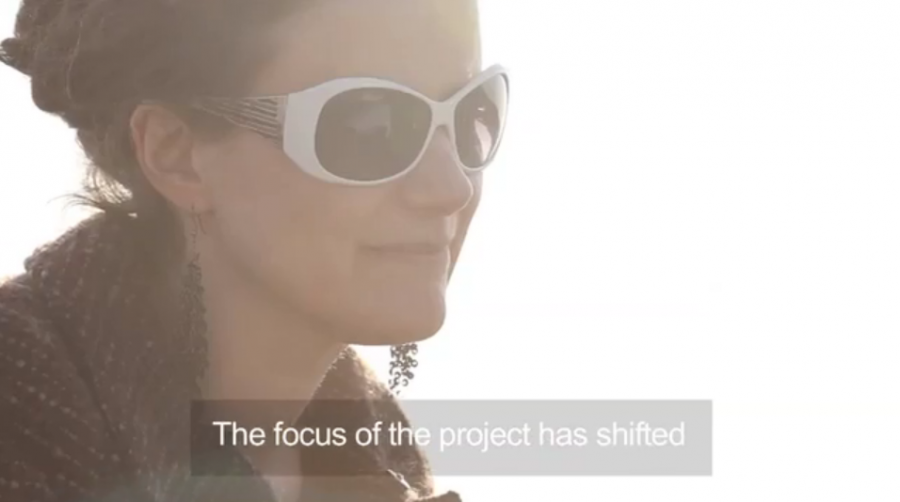 A screen grab of a captioned film. There is a woman's face, shot against the sun, she is wearing long earrings and sunglasses. The text says 'The focus of the project has shifted'