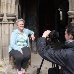 Bobby Baker sitting in the sunshine by a church entrance arch being filmed by a camera man