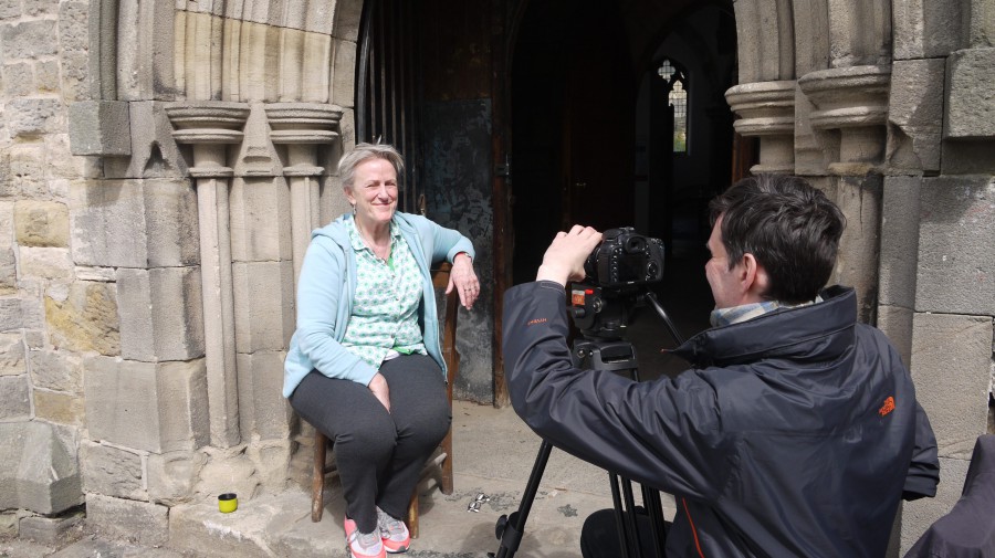 Bobby Baker sitting in the sunshine by a church entrance arch being filmed by a camera man