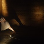 Claire Cunningham dancing with her crutches in a cellar with deep shadows. She is in a simple beige shift , one crutch is on the ground and she has her weight on the other with her body off to the right.