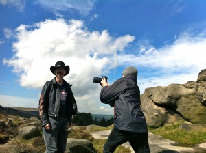 K+Jex Colborne, in a leather jacket and stetson, is being filmed by a camera person against a dramatic backdrop of Yorkshire moors and cloud filled blue sky