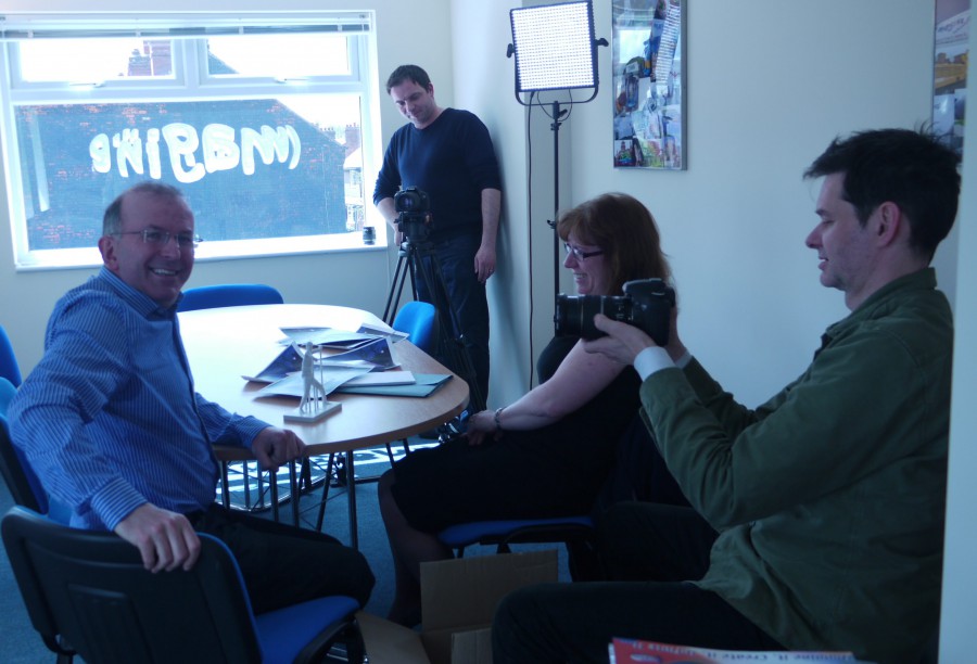 Simon sits at a table, smiling at the camera. He is with a woman and being filmed by two camera men.