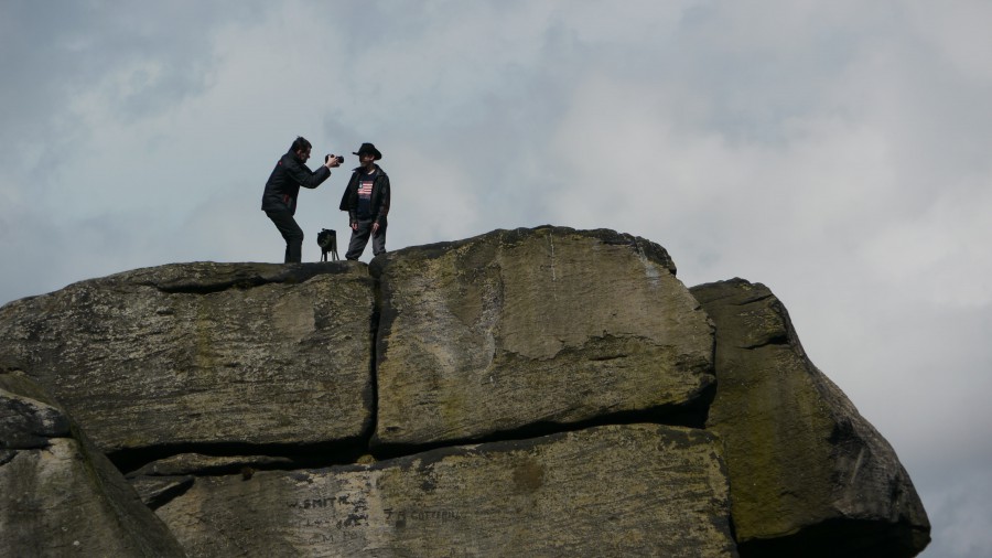 A huge rocky outcrop against a cloudy sky - two figures can be seen - one filming, one in a stetson next to an old air raid siren