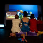 Three performers are watching a screen with their backs to us, they are dressed in circus clothes and are sitting on a fourth performer. The screen they are watching has a playmobil circus on it.