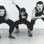 Three white faced mimes, dressed in black, in crouched poses facing the camera