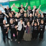 a group of about 20 people, shot from above, all with their arms in the air (and all wearing black 'streetwise choir' tshirts)