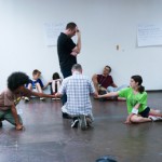 image in a rehearsal room - one figure stands his head in his hand and five others are on the floor around him forming a circle with their arms.