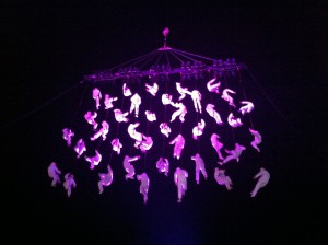 40+ people in white boiler suits are suspended in the air as though on a net. They are lit pink.