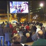 Image of the Plymouth big screen at dusk with a crowd of people gathered to watch the screen showing a band performing.