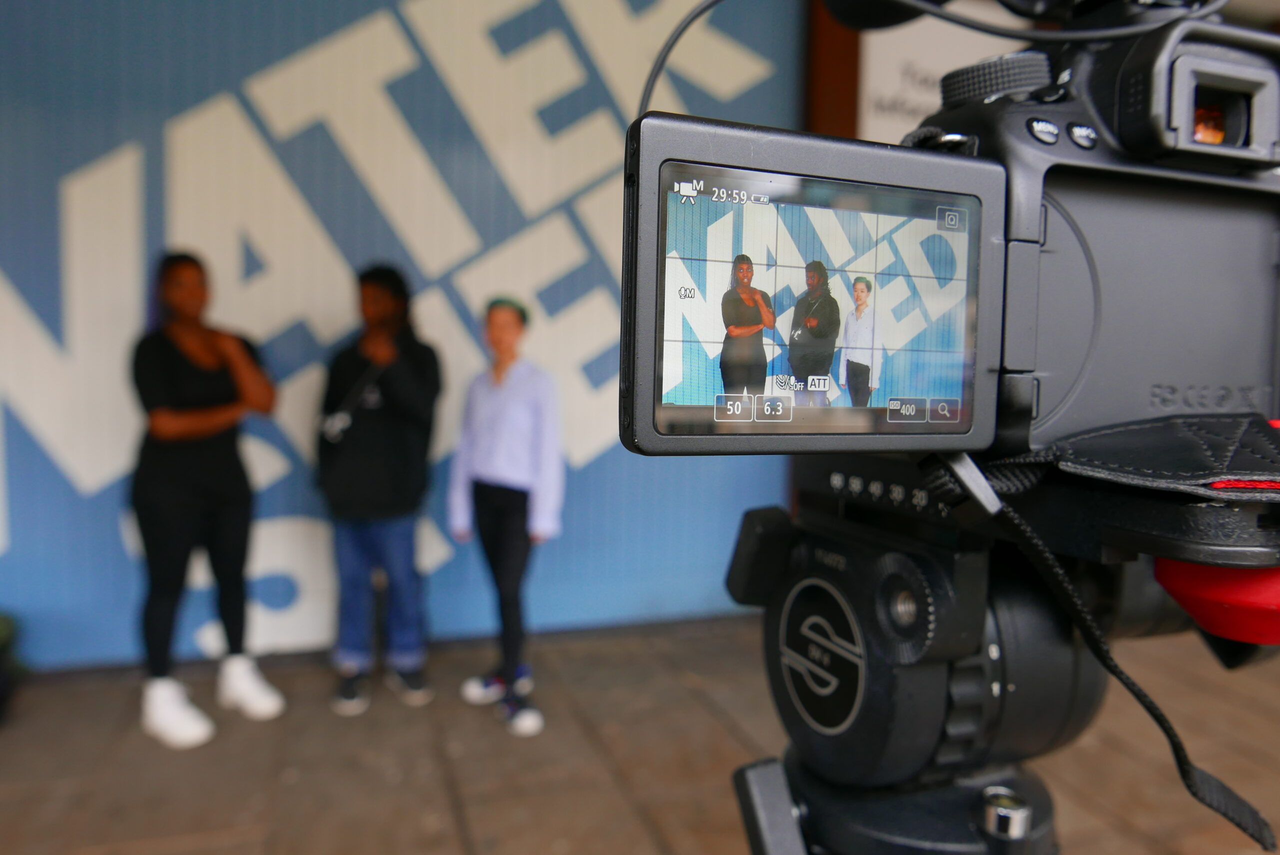 A group of young people posing in front of Watershed frontage. A camera monitor is in the foreground. 