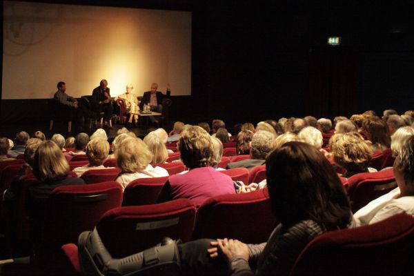 A warmly lit cinema, we see the back of peoples head sat on red chairs. Looking at a few people seated on a stage