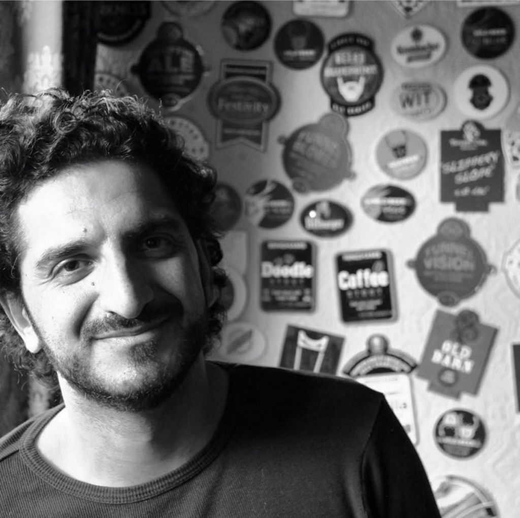 Headshot of Timon Singh. A black and white image of a smiling man in a dark top with short curly hair in front of a wall covered what appear to be beer mats