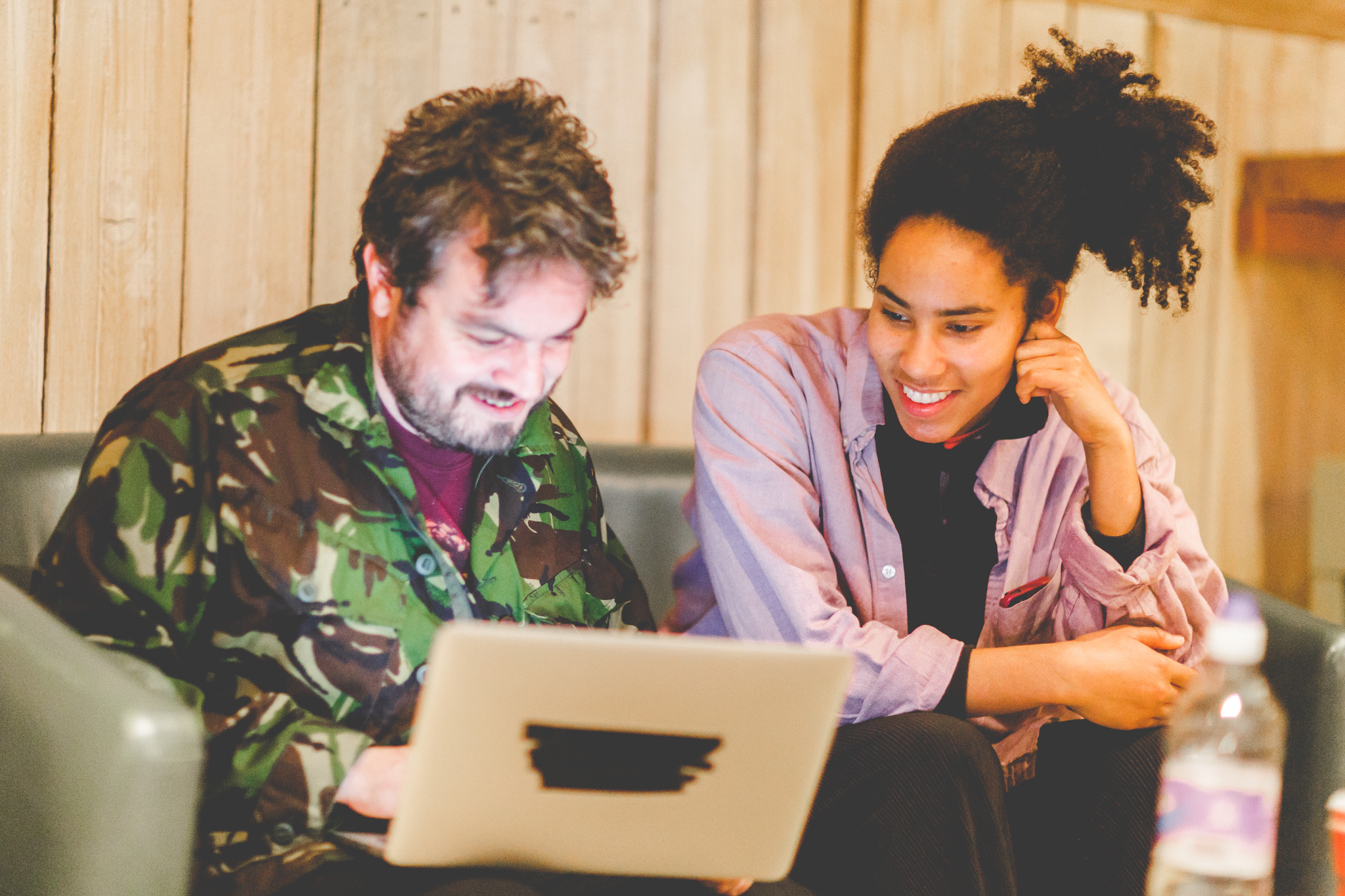 A boy and girl sit on a sofa, smiling at a laptop which illuminates their faces. The boy is dressed in a camo shirt, and the girl a pink one, the room appears warm and cosy in neutral tones.