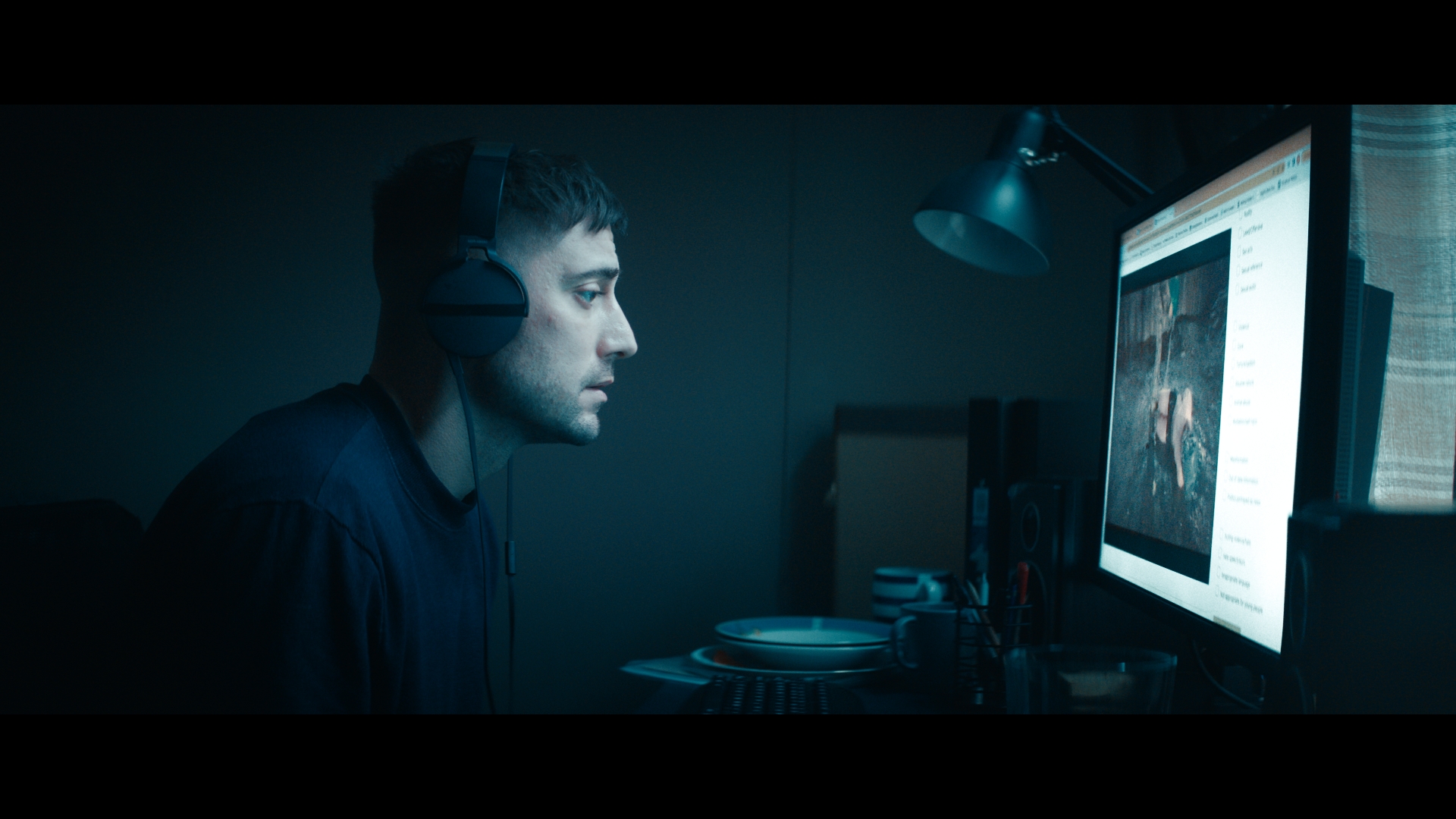 a white man with headphones on stares at a computer screen in the dark