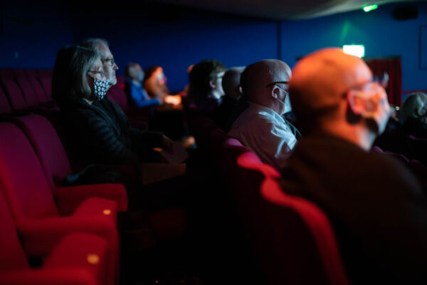 a masked audience sit in an auditorium, they are watching a film, warmly lit by the screen hues of orange and blue