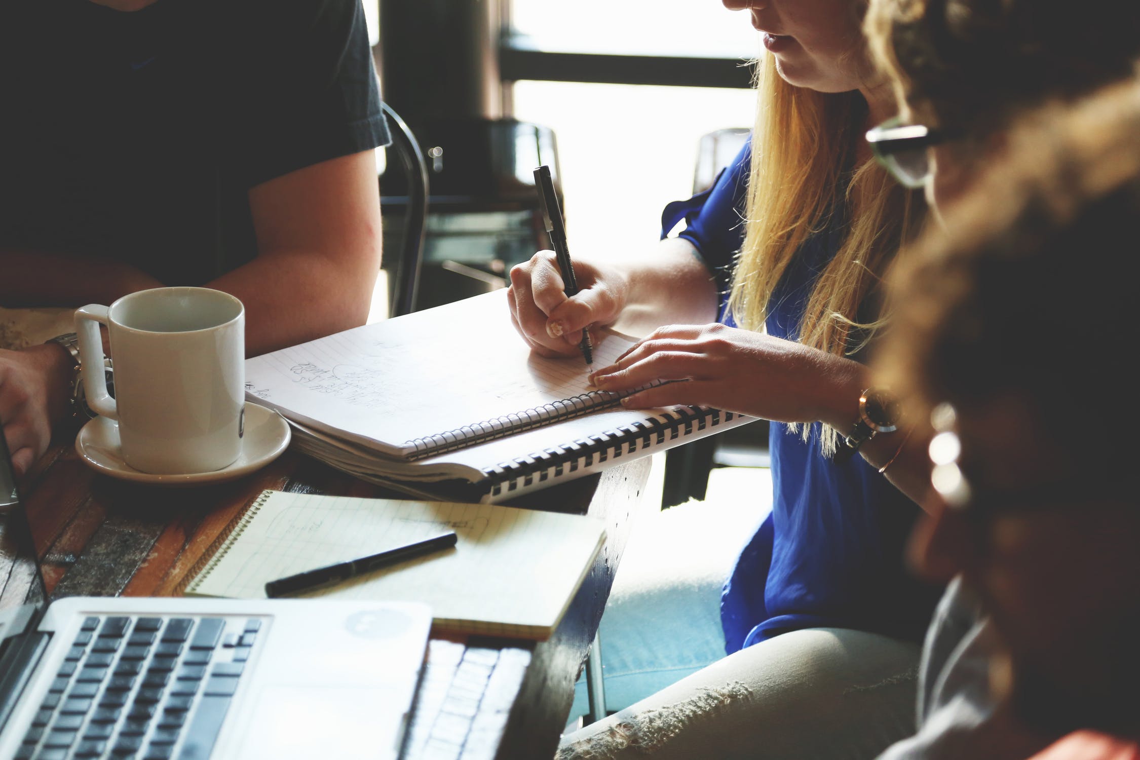 Image of a table, the hands of people around it are visible as they write. There are pads, coffee cups, and a laptop. The image generates a feeling of a lively creative space