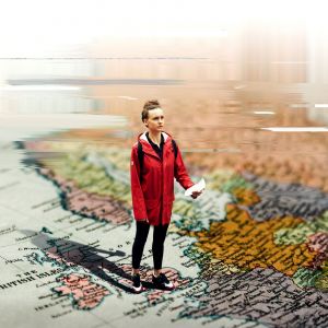 A woman in a red coat stands looking bewildered holding a piece of paper. We look at her standing looking comically large stood on a printed map of the world. However, her posturing and the angle looking down makes her look small.