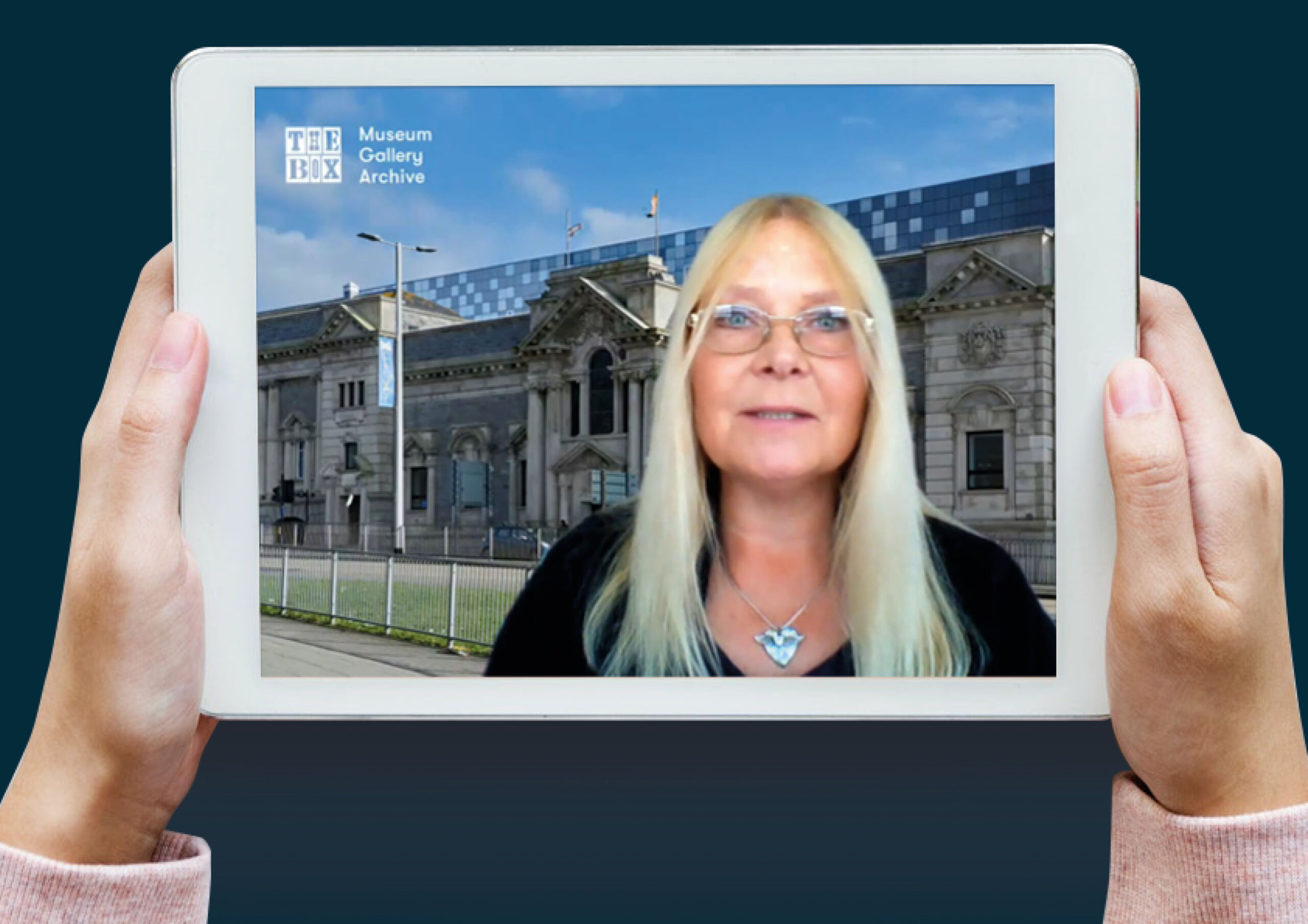 Image of two hands holding an iPad, a white rectangle with an image on. The image is of a woman stood infront of an building, as if presenting.
