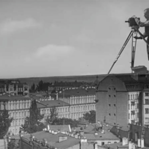 black and white film still. a larger than life person operates a camera, stood as though on the roofs of many houses