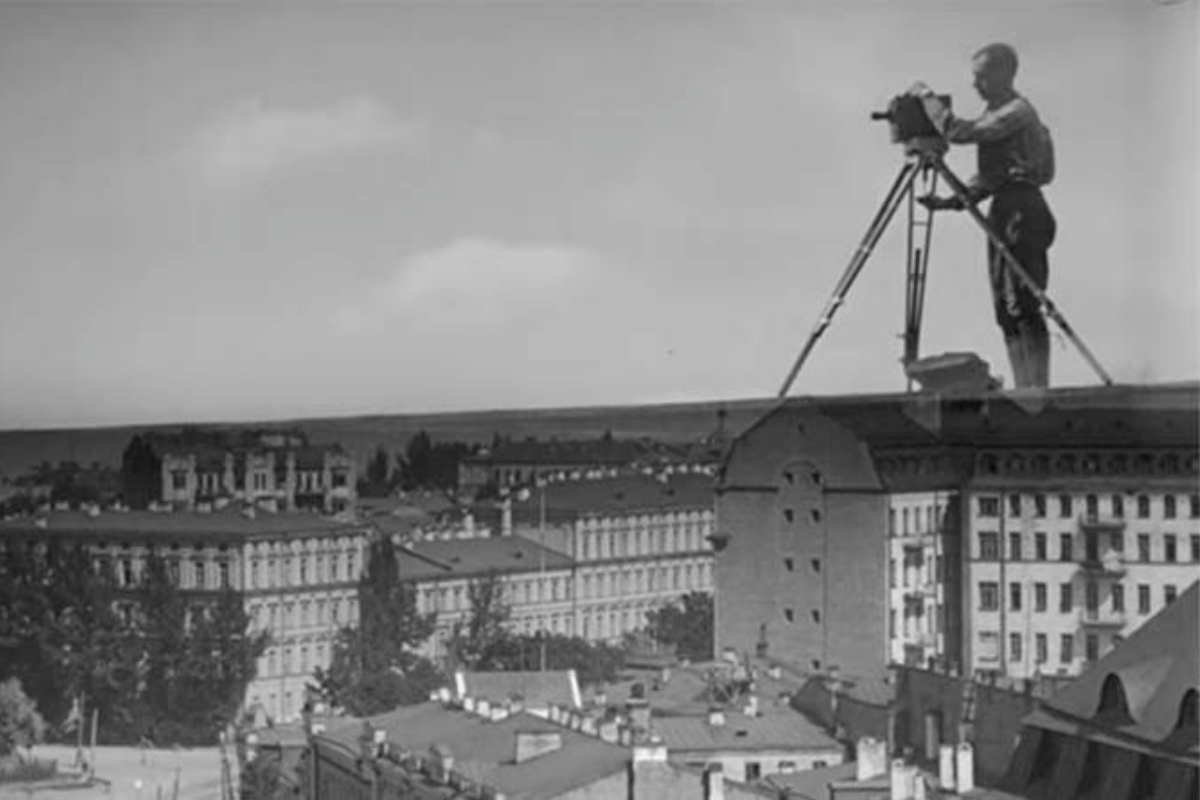 black and white film still. a larger than life person operates a camera, stood as though on the roofs of many houses