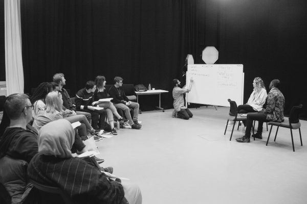 black and white image of a man teaching a workshop. a group of people sit in a curved semi circele, as one draws on a whiteboard