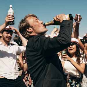 central image of Mads Mikkelsen drinking a bottle of champagne with it in the air - a crowd dressed as sailors cheer him on. the sky is blue, it appears a fun sunny day