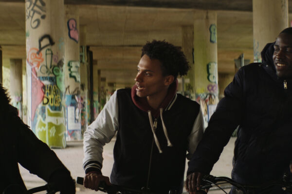 three boys on bikes talk and laugh between each other, riding under a heavily graffitied underpass