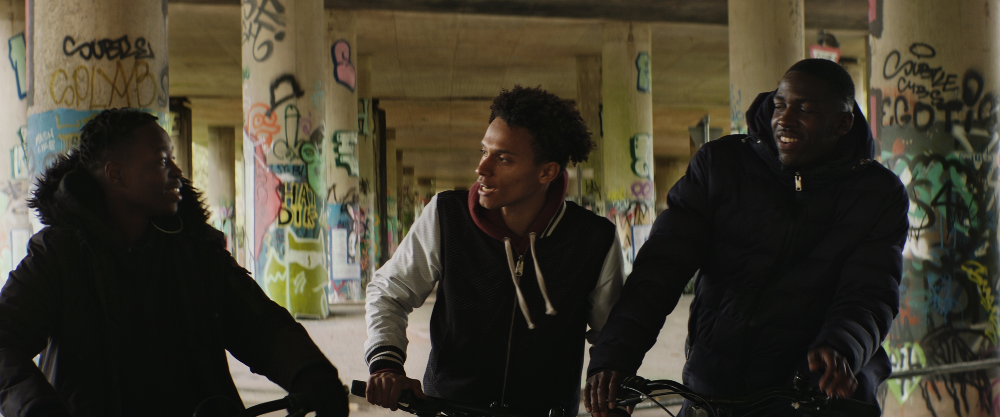 three boys on bikes talk and laugh between each other, riding under a heavily graffitied underpass