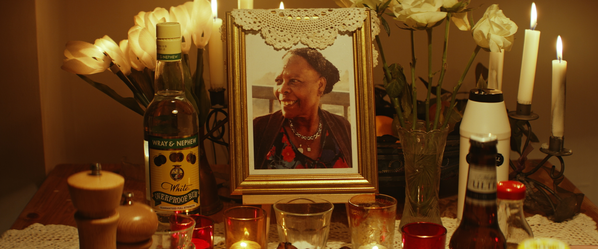 a shrine to a woman at her nine nights. a warmly lit table with candles has a gold framed photo, bottle of rum