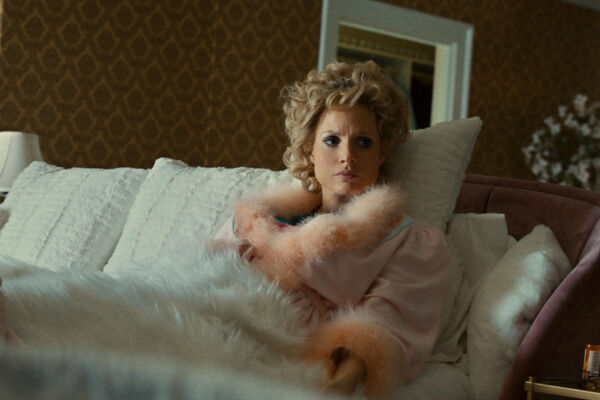 A white woman sat upright in bed. she looks frustrated, and is dressed decadently in feathers with her hair done up.