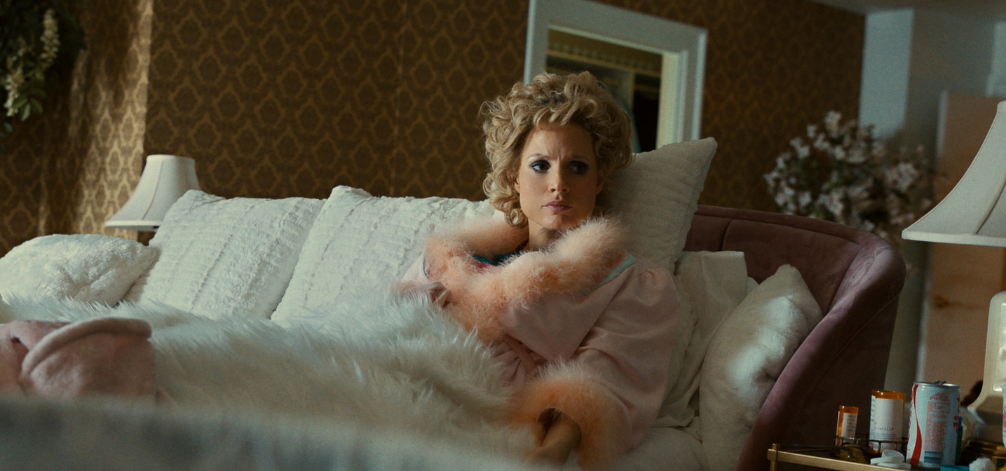 A white woman sat upright in bed. she looks frustrated, and is dressed decadently in feathers with her hair done up.