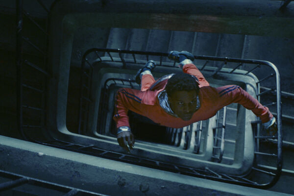 Film still of Gagarine. A boy floats through a spiral stairwell. Suspended in mid air, he wears an orange spacesuit. The stairs are lit a cool teal blue in contrast.