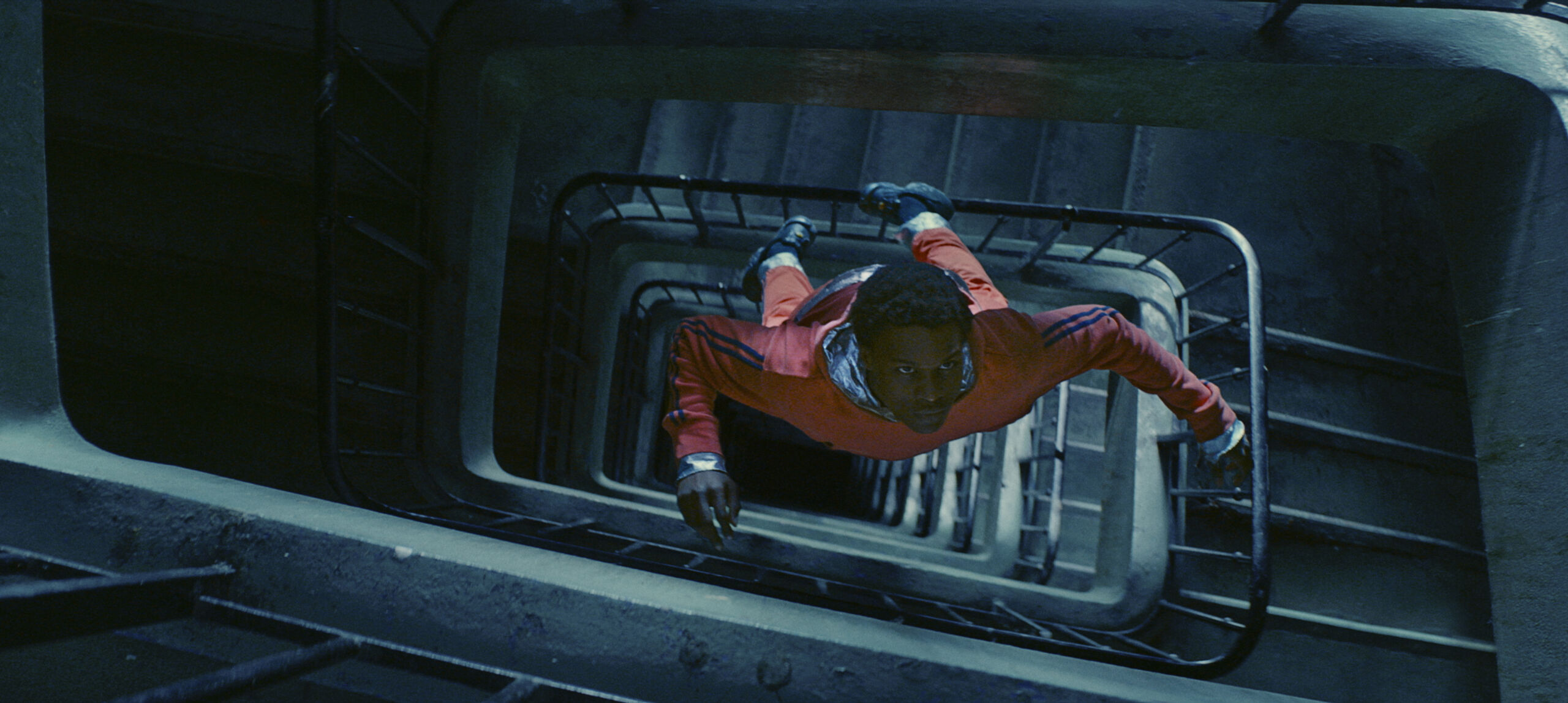 Film still of Gagarine. A boy floats through a spiral stairwell. Suspended in mid air, he wears an orange spacesuit. The stairs are lit a cool teal blue in contrast.