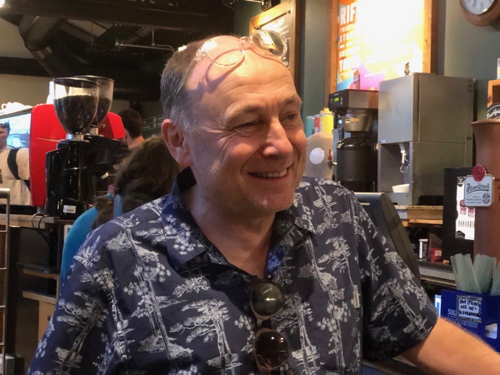 Mark Cosgrove smiles in a printed shirt, stood at the bar. he is a smiling bald white man of middle age