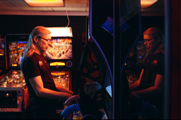 film still from CANNON ARM AND THE ARCADE QUEST. A man plays on a game stand in a dimly lit arcade. he has long white hair, and glasses. the mood is retro and cosy