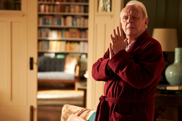 Film Still from THE FATHER. Anthony Hopkins. Film still. an old white haired man stands in a decadent home. he stands holding his hands together in a plush red dressing gown, the room is warmly lit and lined with books
