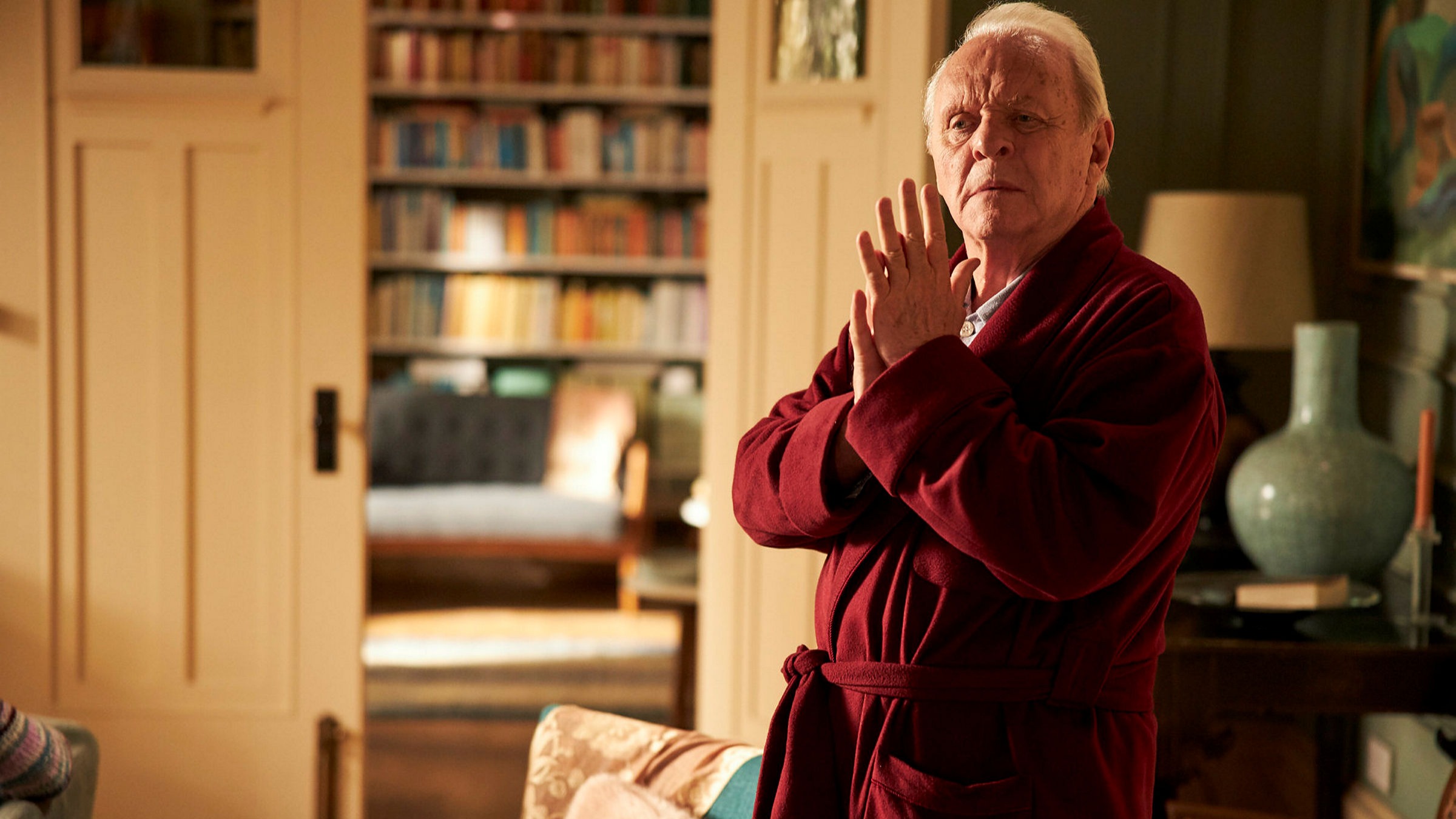 Film Still from THE FATHER. Anthony Hopkins. Film still. an old white haired man stands in a decadent home. he stands holding his hands together in a plush red dressing gown, the room is warmly lit and lined with books