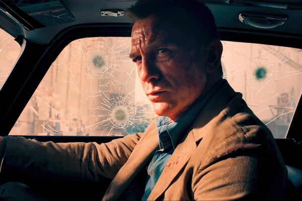 Still from James Bond film NO TIME TO DIE. A moody lit dark shot of bond, played by Daniel Craig, sat in a car. he is a white man and government agent.. he looks serious and pondering. bullet holes mark the glass of the car windows he sits in