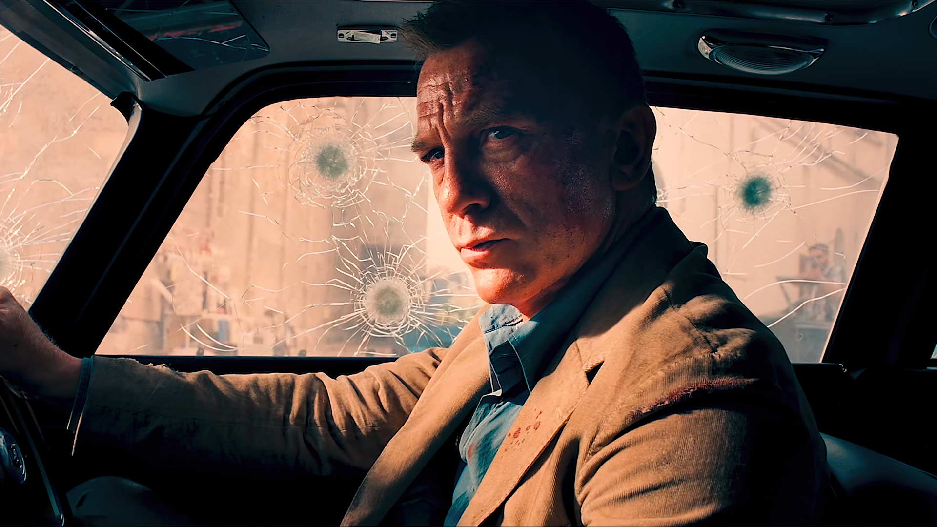 Still from James Bond film NO TIME TO DIE. A moody lit dark shot of bond, played by Daniel Craig, sat in a car. he is a white man and government agent.. he looks serious and pondering. bullet holes mark the glass of the car windows he sits in