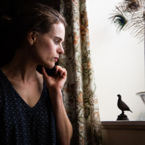 a woman in a darkened room looks out a white window,. she appears concerned and on the phone. a small silhouette of a bird state stands out against the white wall