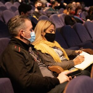 Two people sit in a cinema. they are masked, and taking notes. The cinema is well lit for a conference event, showing comfy puple seats. The two people are centre, one taking notes, the other listening intently.