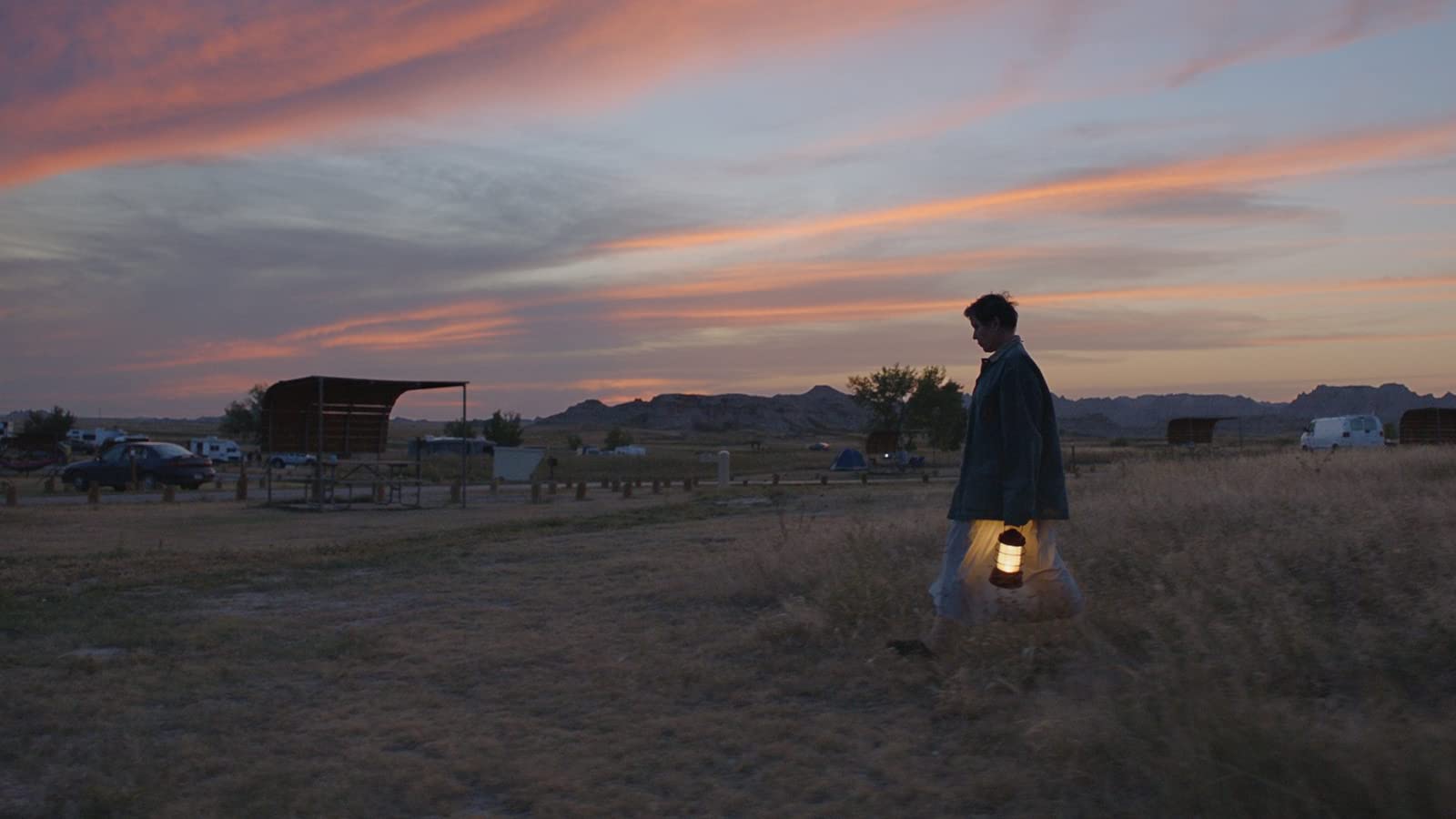 a dusk scene of a setting sun, pink Sky over the desert. a single woman walks towards a faraway caravan encampment. she carries a warmly lit torch